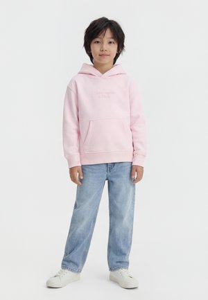 Copil stând îmbrăcat într-un hanorac roz deschis, blugi albastru deschis și adidași albi pe un fundal alb simplu.