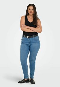 Vrouw met gekruiste armen, gekleed in een zwart mouwloos shirt, blauwe jeans, een gestikte zwarte riem en zwarte loafers tegen een effen achtergrond.