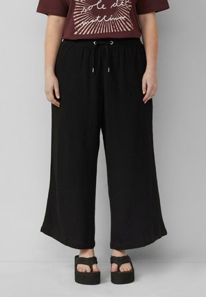 Osoba nosząca luźne czarne spodnie z szerokimi nogawkami, z elastycznym pasem i sznurkiem, w połączeniu z czarnymi sandałami na platformie i bordowym t-shirtem z nadrukiem.