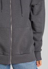 Sudadera con cremallera gris hecha de una tela suave, que presenta una cremallera de longitud completa, puños acanalados y un bolsillo en la parte frontal.