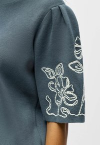 T-shirt bleu en tricot à manches courtes avec épaules bouffantes et motifs floraux brodés blancs sur la manche.