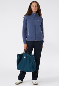 Giacca blu con zip e colletto alto, due tasche laterali; abbinata a pantaloni scuri e una grande borsa teal con manici marroni e borchie.