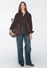 Camicia nera a bottone, jeans blu a gamba larga, scarpe marroni e una borsa in suede scuro con dettagli metallici. Il modello si trova di fronte a uno sfondo bianco.