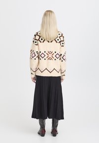Suéter de punto color crema con patrones geométricos en marrón, rosa y negro, combinado con una falda negra con pliegues y botas hasta la rodilla en negro y burdeos.