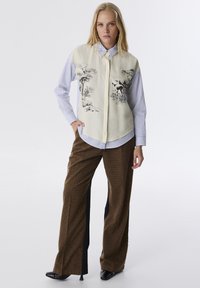 Camicia a strati con una parte anteriore crema e una stampa paesaggistica nera su una camicia a righe blu e bianche. Abbinata a pantaloni a quadri marroni.
