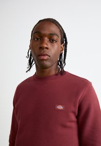 Bordeaux sweatshirt met een ronde hals, voorzien van een klein geborduurd logo-patch op de linkerborst. Glad textuur en een getailleerd ontwerp.