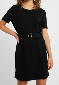 Czarna sukienka z krótkimi rękawami, okrągłym dekoltem i dopasowanym stanem. Posiada szeroki pasek z prostokątną klamrą. Gładki, strukturalny materiał.