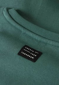 Grön sweatshirt med en svart etikett som står "SKAPA DIN FRAMTID." i vita bokstäver, visar tygtextur och sömnadsdetaljer.