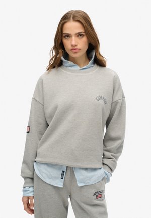 Felpa grigia a maniche lunghe con scollo rotondo, caratterizzata dal logo "Superdry" e una camicia a righe blu sotto, abbinata a pantaloni coordinati.