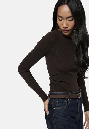 Mujer con cabello largo y oscuro que lleva una camiseta marrón ajustada de manga larga con textura de canalé, metida dentro de unos vaqueros azul oscuro con cinturón marrón, mirando hacia abajo.