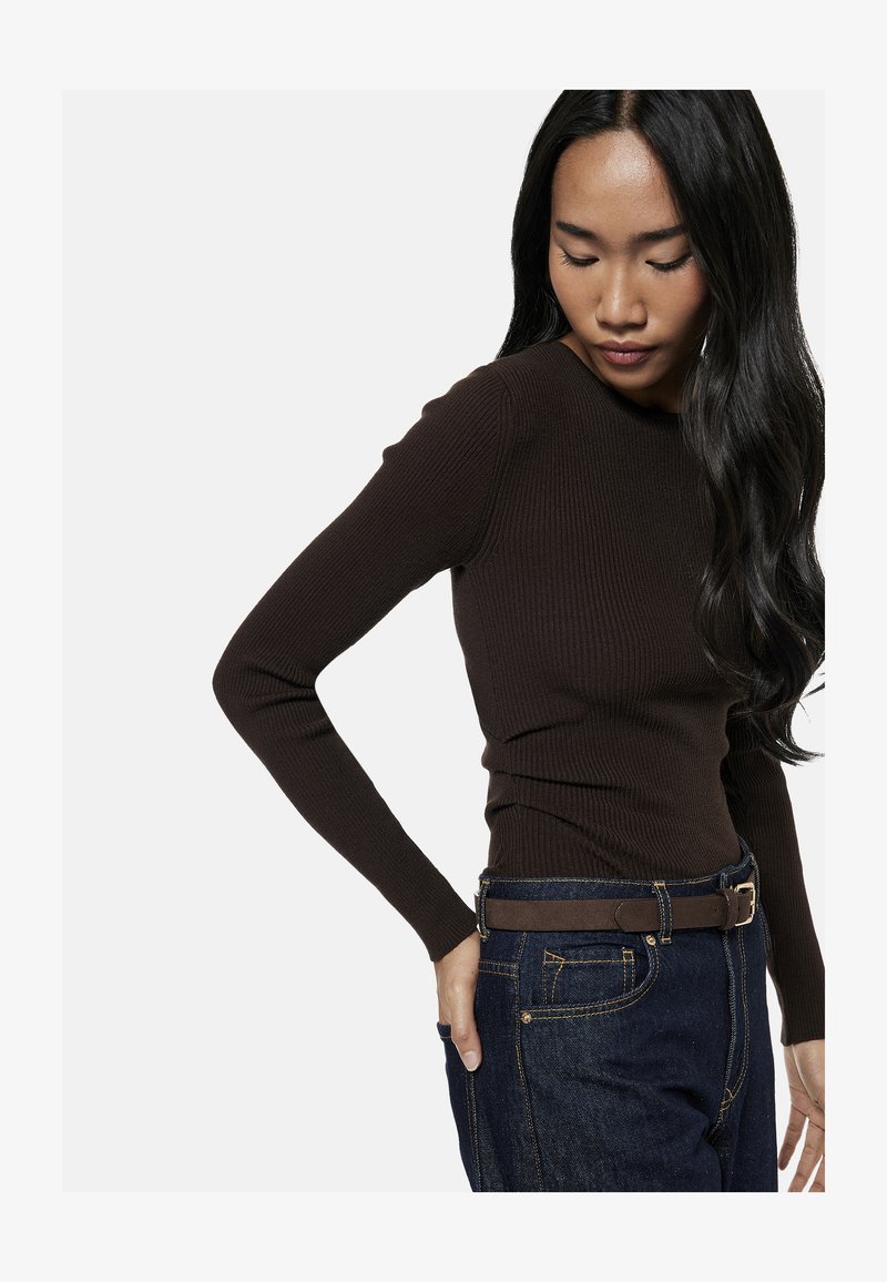 Femme aux longs cheveux foncés portant un t-shirt marron côtelé moulant à manches longues, rentré dans un jean bleu foncé avec une ceinture marron, regardant vers le bas.