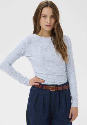 Žena s dlhými hnedými vlasmi, oblečená v svetlomodrom čipkovanom top s dlhým rukávom, zastrčenom do tmavomodrých nohavíc s vysokým pásom a hnedým opaskom.