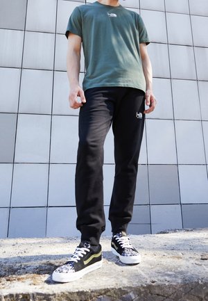 Camiseta de algodón verde, pantalones deportivos negros con logo y zapatillas negras/blancas con patrón floral y franja verde, frente a una pared metálica.