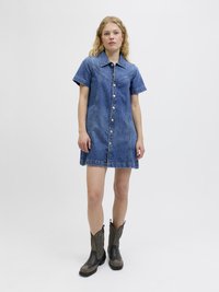 Abito camicia in denim con maniche corte, chiusura a bottoni e colletto strutturato. Colore blu scuro con cuciture visibili. Indossato con stivali cowboy marroni.
