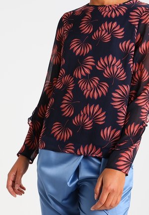 Mørkeblå bluse med rødt palmebladsmønster båret sammen med lyseblå satinbukser, viser lange ærmer og afslappet pasform.