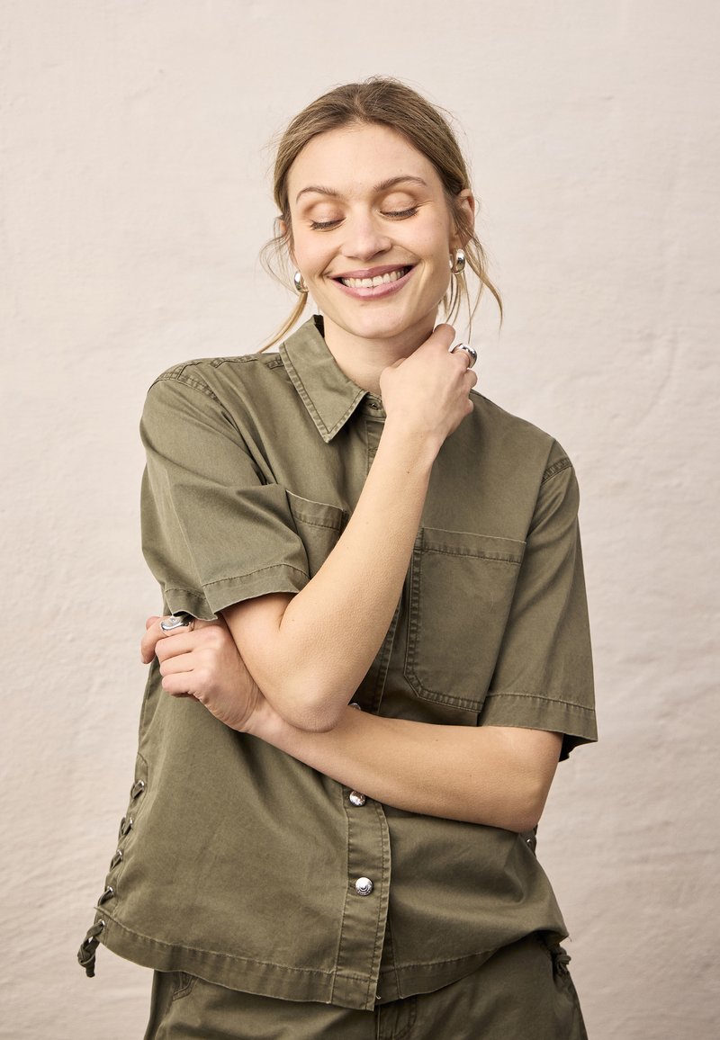 Femme souriante aux yeux fermés portant une chemise boutonnée à manches courtes couleur vert olive, tenant son bras de la main près du visage, debout devant un fond uni.
