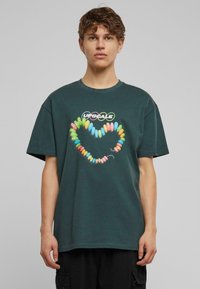 Mørkegrøn t-shirt i bomuld med korte ærmer, der har et farverigt hjerte lavet af candy-lignende former, med teksten "UPSCALE" ovenfor.