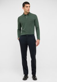 Dunkelgrünes Poloshirt mit langen Ärmeln, Kragen und einer Leiste mit drei Knöpfen, kombiniert mit marineblauen, schmal geschnittenen Hosen und schwarzen Schuhen.