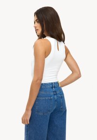 Haut blanc sans manches avec une ouverture en forme de goutte dans le dos, associé à un jean en denim bleu taille haute. Tissu lisse, coupe décontractée et design minimaliste.