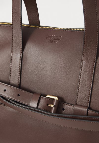 Smeđa kožna torba Moschino Milano s zlatnim patentnim zatvaračem i detaljem kopče, prikazuje izbliza ručke i pojasni remen.