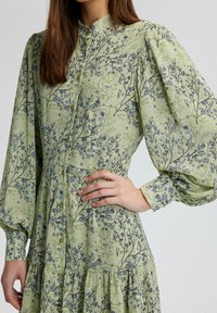 Rochie cu mâneci lungi și nasturi, dintr-un material de culoare verde deschis, ce prezintă modele florale în nuanțe de albastru și negru, cu fustă în straturi și mâneci bufante.