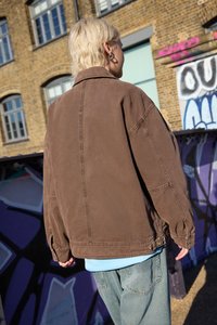 Osoba s krátkymi blond vlasmi oblečená v hnedom saku a modrých džínsach stojí pri múre pokrytým graffitmi v mestskom prostredí.