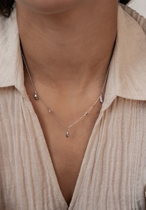 Silver chain necklace with small teardrop pendants worn on a person with light beige textured shirt and open collar.