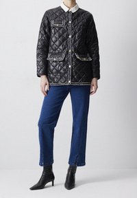 Person iført en sort quiltet jakke med guldknapper og trim, blå jeans med ankel-splits og sorte spidsede støvler.