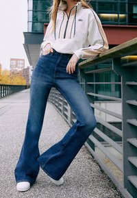 Bílá mikina na zip s černými a béžovými akcenty; modré džíny s vysokým pasem a rozšířenými nohavicemi; bílé tenisky; městské venkovní prostředí.
