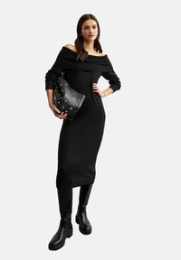 Svart off-shoulder stickad klänning med ribbad halsringning och långa ärmar, tillsammans med en svart väska med silverringdetaljer och knähöga stövlar.