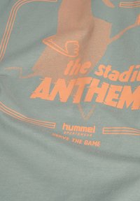 Ljusgrön t-shirt med orange tryckt text och grafisk design. Har logotyperna "the stadium ANTHEM" och "hummel SPORTSWear". Mjuk tygtextur.