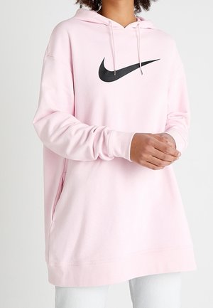 Osoba nosząca jasnoróżową bluzę z kapturem Nike z czarnym logo swoosh, złożone ręce, w zestawieniu z jasnymi spodniami.