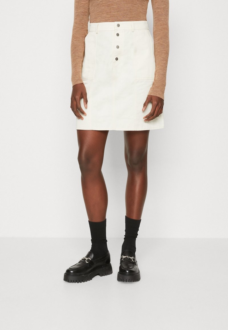 Personne portant une jupe blanche boutonnée à l'avant, un haut marron à manches longues, des chaussettes noires et des mocassins noirs épais sur un fond uni.