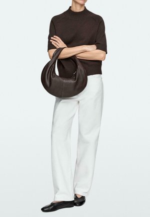 Persoon draagt een donkerbruin kort mouwensweater en witte broek, en houdt een grote donkerbruine leren handtas in de vorm van een halve maan vast.