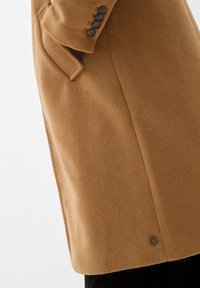 Cappotto di lana color cammello con tasche a patta e quattro bottoni scuri sui polsini, indossato sopra pantaloni neri.