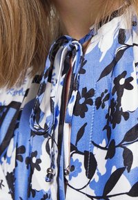 Blauw-witte blouse met bloemenprint en een striksluiting. Voorzien van een ronde hals, korte mouwen en opvallende zwarte bloemenmotieven. Soepele textuur.