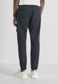 Man in zwart tapse cargobroek met elastische boorden en witte sneakers, staand tegen een effen lichte achtergrond.