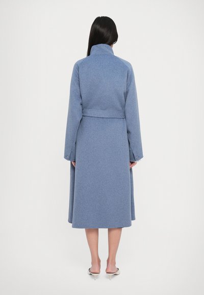 Femme aux longs cheveux foncés portant un manteau bleu clair ceinturé et des mules blanches à talons, se tenant de dos sur un fond blanc uni.