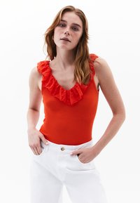 Body rosso a coste con scollatura in pizzo volant. Abbinato a pantaloni in denim bianchi a vita alta. La modella si trova con le mani sui fianchi davanti a uno sfondo neutro.