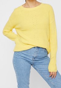 Kvinna i en lös gul stickad tröja med subtila mönster och ljusblå högmidjade jeans, poserande med handen på höften.