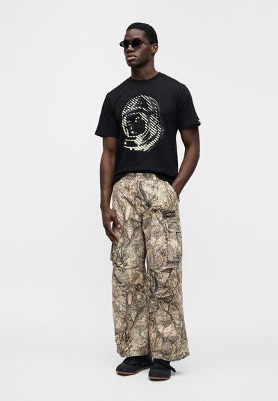 Μαύρο μπλουζάκι με γραφική εκτύπωση προσώπου αστροναύτη, συνδυασμένο με φαρδιά παντελόνια καμουφλάζ τύπου cargo και μαύρα παπούτσια.