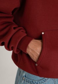 En persons hånd i lommen på en rød sweatshirt med manchetter, iført lyseblå denimjeans.