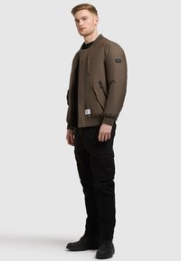 Veste bomber marron avec des poignets côtelés, des poches zippées sur le côté et un patch logo. Associée à un pantalon cargo noir et des bottes noires.