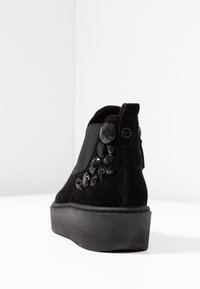 Černá semišová kotníková bota s elastickým bočním panem, platformovou podešví a dekoračními černými drahokamy na vnější straně.
