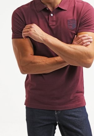 T-shirt polo bordeaux avec un col classique, deux boutons et un petit logo brodé sur la poitrine gauche, associé à un jean en denim foncé.