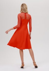Robe rouge avec un corsage ajusté, des manches transparentes à pois et une jupe évasée. Le dos présente une fermeture éclair dissimulée et un détail texturé. Talons transparents.