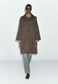 Cappotto oversized marrone in tessuto testurizzato, con colletto ampio, tre bottoni e maniche larghe, indossato sopra jeans chiari.
