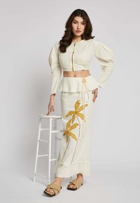 Cream cropped jacket with puff sleeves, zip closure; matching long skirt with yellow floral embroidery and frayed hem. Beige sandals.