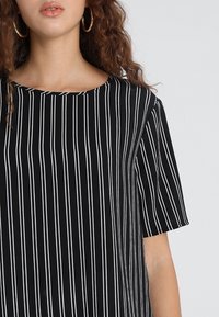 Maglietta a righe bianco e nero con maniche corte, colletto rotondo e una trama di tessuto liscia. Presenta righe verticali di larghezze variabili.