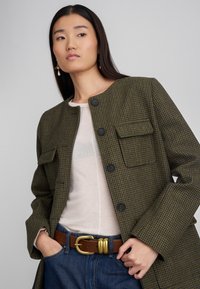 Zielono-brązowy płaszcz w kratę z okrągłym kołnierzem, czarnymi guzikami i dwiema przednimi kieszeniami, zestawiony z beżową bluzką i niebieskimi dżinsami.